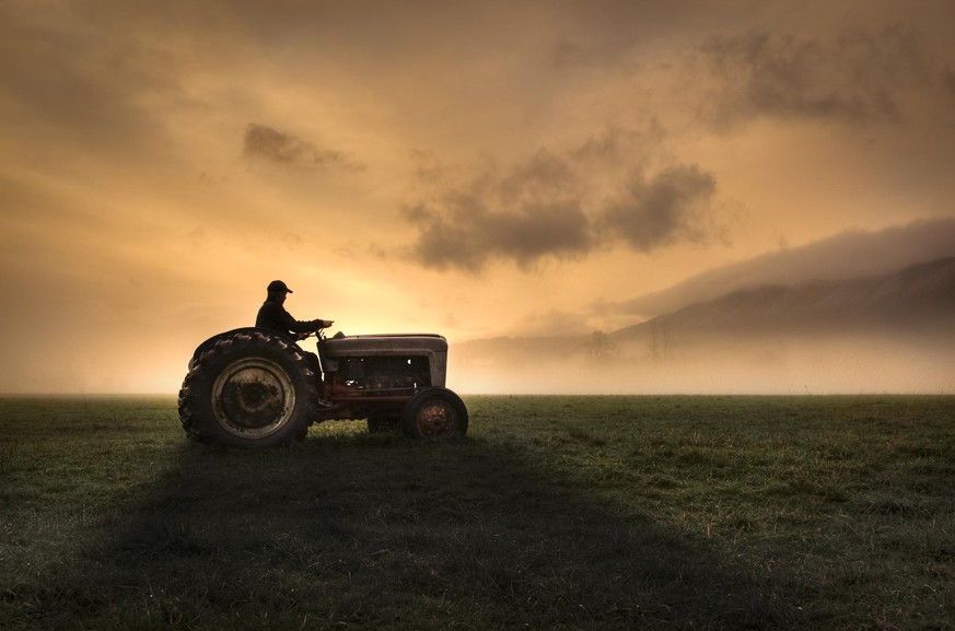 Farmer riding tractor during sunrise with vast open fields and bright orange sky on chilly, foggy morning.
