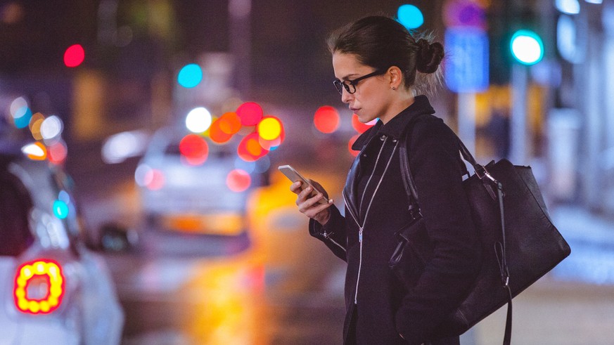 Close-up of a young woman using smart phone on the busy city street at night. The woman is serious while looking at the phone. She is dressed in black coat and wears a big black purse. Her long dark h ...