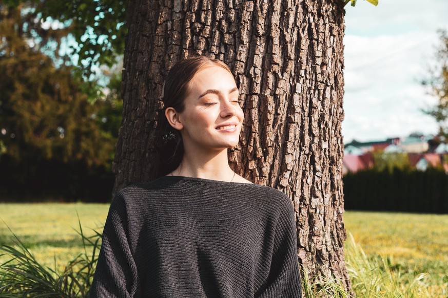 Young woman leaning on tree enjoying the warm sunlight. Enjoying the silence, smiling happy.
