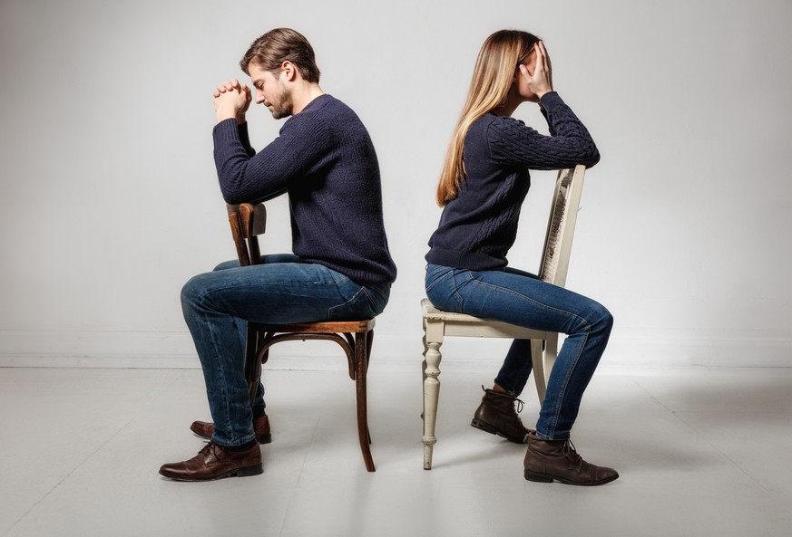 Side view of depressed couple sitting back to back on chairs against gray background