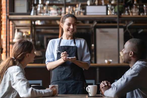Attractive waitress laughing at African American man funny joke, serving customers, diverse couple making order in cafe, coffeehouse female worker and multiracial visitors having fun