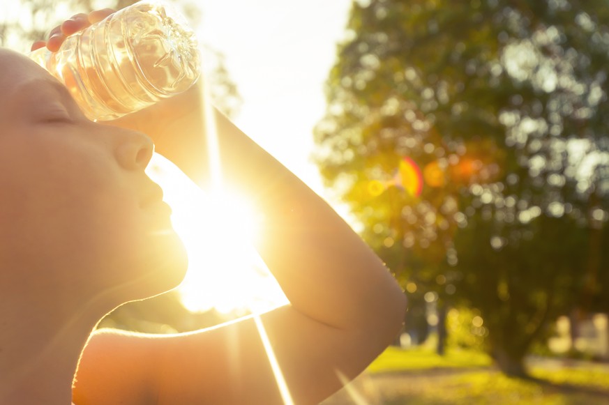 Woman using water bottle to cool down. Fitness and wellbeing concept with female athlete cooling down on a city street. She is holding a water bottle to her head to cool down. The sun is low creating  ...