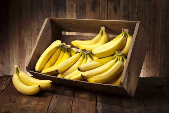 Side view of a tilted wooden crate filled with fresh organic bananas sitting on rustic wood table. The crate is tilted with some bananas out of it. Predominant color is brown and yellow.Low key DSRL s ...