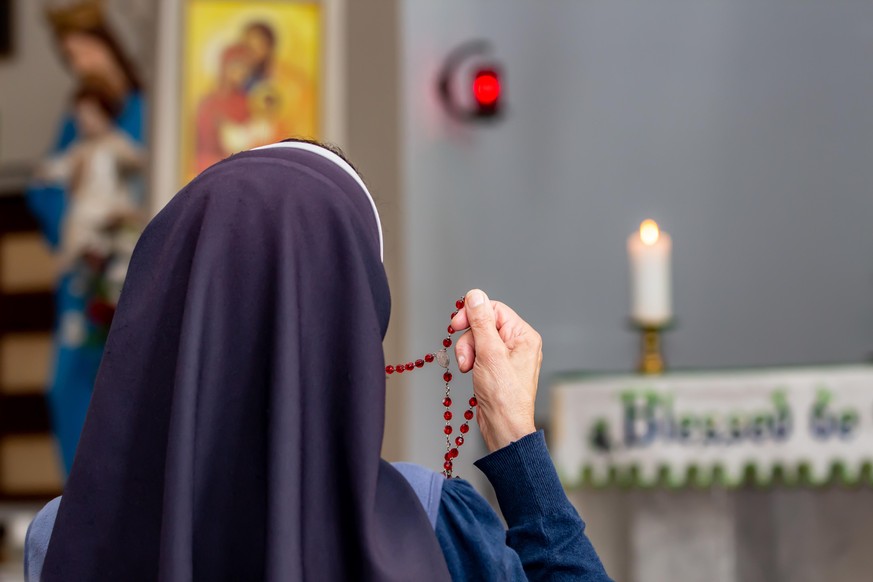 View from back of a religious sister holding rosary and praying at altar in church chapel.