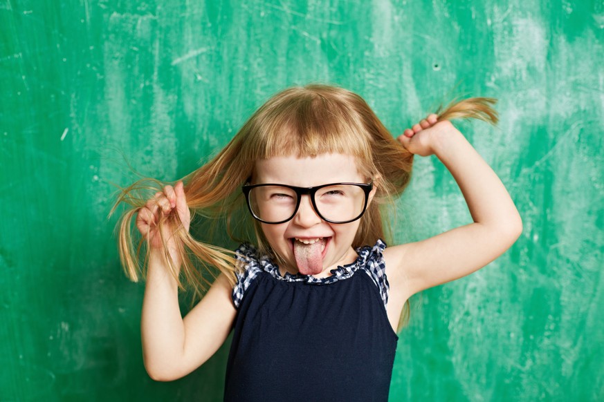Little child with mischief looking at camera. Cheerful brat holding hair and sticking tongue out. Cute girl wearing eyeglasses. Its photo illustrating childhood. It is perfect for using it in commerci ...