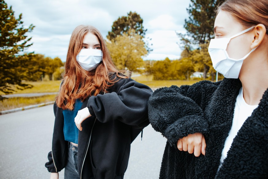 Two Teenage Girls doing an Elbow Bump to greet each other meeting outside during Covid-19 pandemic times. Wearing surgical face masks greeting each other securely with an elbow bump. Avoiding tight bo ...