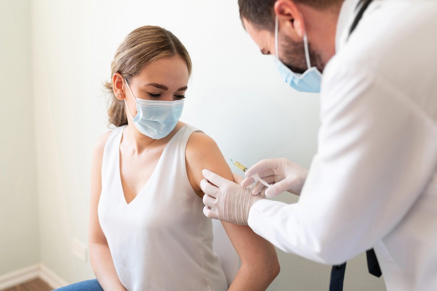 Closeup of a nervous woman and her doctor wearing face masks and getting a vaccine shot in a doctor&#039;s office